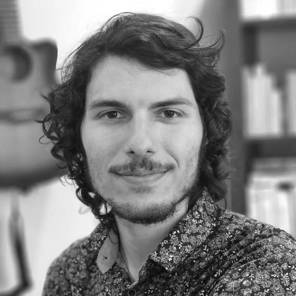 Photo de profil en noir et blanc du concepteur web Xavier Harel, jeune homme aux yeux foncés, ayant cheveux foncés mi-long ondulés, moustache et barbe légère, portant une chemise fleurie, avec en arrière-plan une guitare acoustique et une bibliothèque de livres.