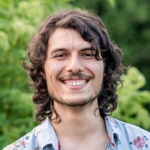 Photo de profil du concepteur web Xavier Harel, jeune homme aux yeux foncés, ayant cheveux foncés mi-long ondulés, moustache et barbe légère, portant une chemise fleurie, avec en arrière-plan feuilles d'arbre.